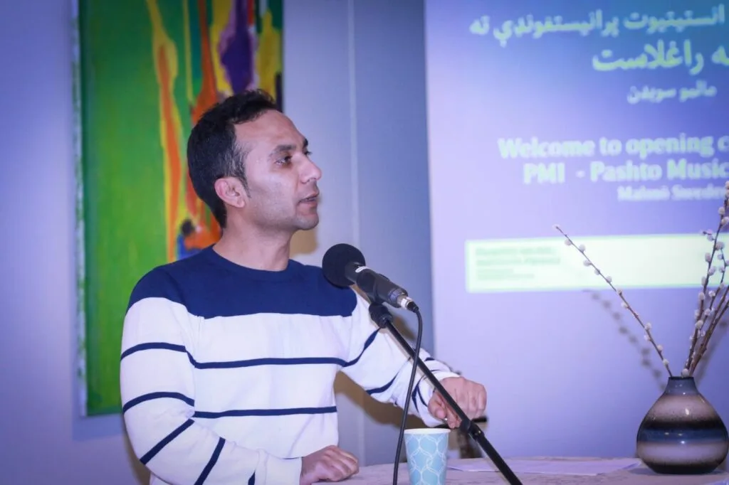 Pashto Music Institute Launches in Malmö, Sweden, Heralding a New Era of Musical Heritage Preservation Saeed Shinwari, an Afghan journalist and poet, speaking at the podium during the Pashto Music Institute's launch. A colorful abstract painting and a presentation screen are visible in the background.