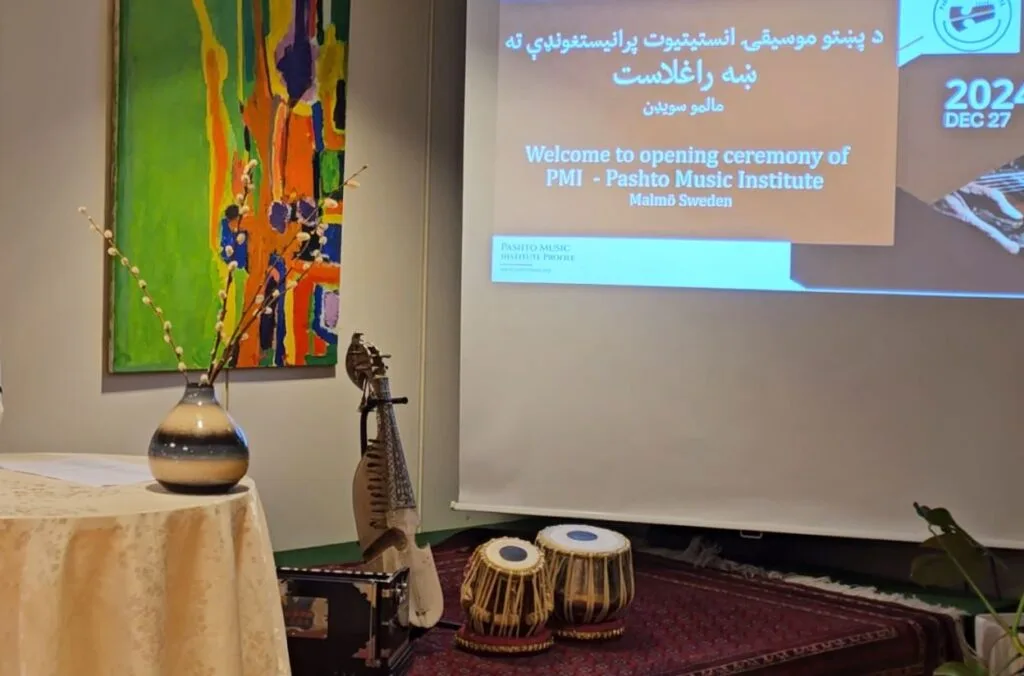 Pashto Music Institute Launches in Malmö, Sweden, Heralding a New Era of Musical Heritage Preservation A photo of the stage at the Pashto Music Institute launch event in Malmö, Sweden, featuring traditional Pashto instruments (rubab and tabla) placed on a red carpet, a decorative vase on a table, and a colorful abstract painting in the background. A presentation screen displays the event's welcome message and details in Pashto and English.