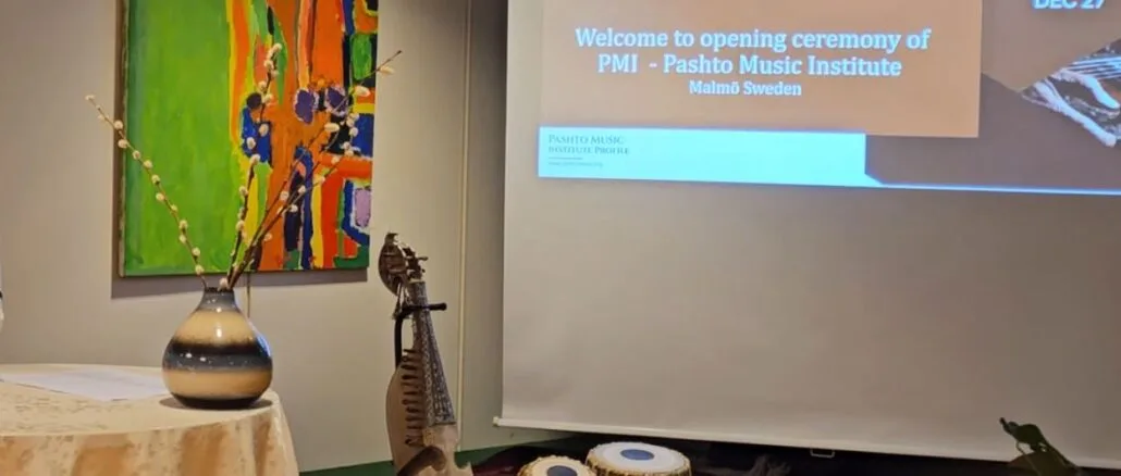 Capture A photo of the stage at the Pashto Music Institute launch event in Malmö, Sweden, featuring traditional Pashto instruments (rubab and tabla) placed on a red carpet, a decorative vase on a table, and a colorful abstract painting in the background. A presentation screen displays the event's welcome message and details in Pashto and English.