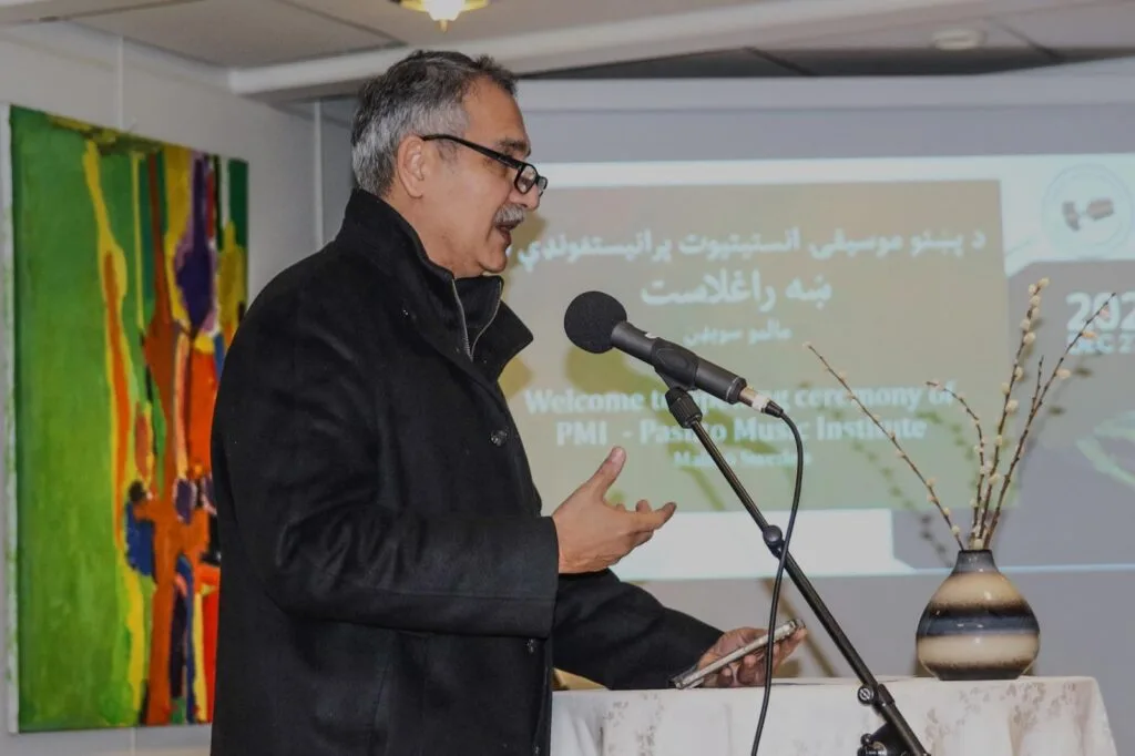 Pashto Music Institute Launches in Malmö, Sweden, Heralding a New Era of Musical Heritage Preservation Haroon Bacha, a prominent Pashto singer and Chairman of the Pashto Music Institute, speaking at a podium during the institute's soft launch in Malmö, Sweden. He is dressed in a black coat, holding a phone in one hand, and speaking into a microphone. A colorful abstract painting and a vase with decorative branches are visible in the background. A presentation screen displays the event details in Pashto and English.