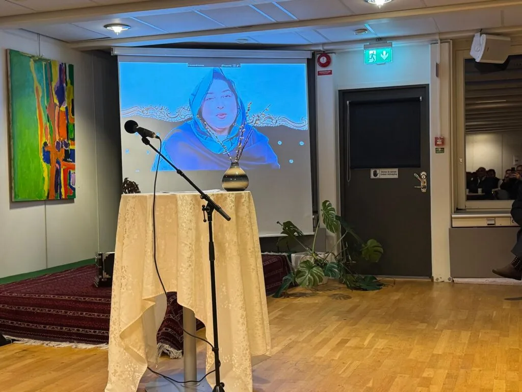 Pashto Music Institute Launches in Malmö, Sweden, Heralding a New Era of Musical Heritage Preservation A photo of a screen showing a video message from Shakila Naz, a prominent Pashto singer, during the Pashto Music Institute’s soft launch event in Malmö, Sweden. The stage setup includes a decorated podium with a microphone, a vase, and traditional Pashto elements.