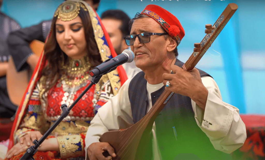 Afghan Musicians Silenced: The Struggle for Survival Under Taliban Rule Mir Muftoon, a renowned Badakhshan folk singer, wearing traditional attire and performing on stage with a rubab, accompanied by a female artist in colorful Afghan dress.