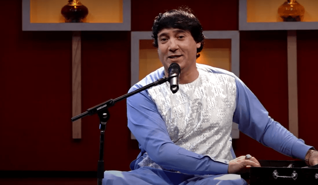 Afghan Musicians Silenced: The Struggle for Survival Under Taliban Rule Nasir Parwani, a well-known Dari-language singer, seated on stage in traditional Afghan clothing, singing into a microphone during a live TV concert in Kabul.