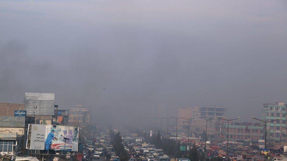 A busy street in Kabul, Afghanistan, covered in dense gray smog. Traffic, billboards, and buildings are barely visible due to the pollution, highlighting environmental and health challenges.