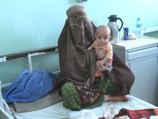 A woman wearing a brown burqa sits on a hospital bed holding a malnourished infant, with hospital items on a nearby table.