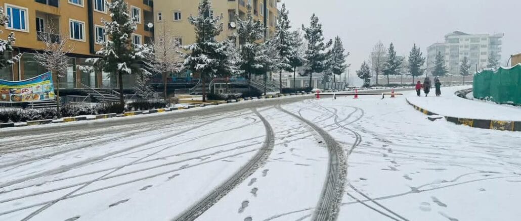 Snowfall in Kabul A snowy street in a Kabul neighborhood with apartment buildings on one side, pine trees covered in snow, and footprints and tire tracks visible on the snow-covered road.