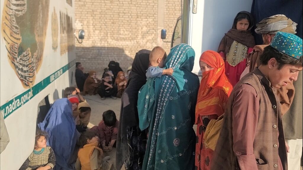 A group of Afghan refugees, including women in colorful traditional attire and children, stand and sit near a large vehicle at the Chaman border crossing on October 31, 2023. Some children sit on the ground, while others cling to their mothers, reflecting the difficult journey ahead.