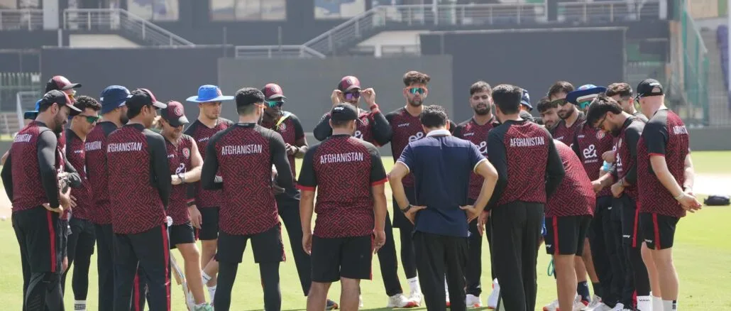 Afghanistan_Cricket_Team_Training_Champions_Trophy_2025_ACB A group of Afghanistan cricket players wearing maroon and black training jerseys and pants with 'Afghanistan' written on the back, standing in a tight huddle on a green field at the National Stadium in Karachi. The players, wearing caps, sunglasses, and athletic shoes, listen intently to a coach or official in a dark blue shirt standing in the center. Cricket bats, water bottles, and training equipment are visible on the grass. The background shows stadium seating, a black wall, and logos for 'Etisalat' and 'vivo.' The image includes 'ACB MEDIA' and 'vivo' branding in the corners, with sunlight casting shadows on the field.