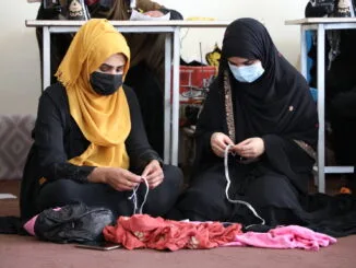 The Cost of Isolation: Afghanistan’s Financial Challenges and Development Prospects Two Afghan women wearing black clothing and headscarves, one with a yellow scarf, sit on the floor of a Women Empowerment Center in Kabul, weaving fabric and string. They are engaged in skill development activities, with sewing machines and other workers visible in the background.
